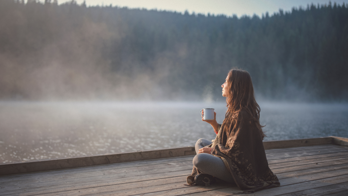 Woman sitting on a dock by a lake, holding a mug, with misty background