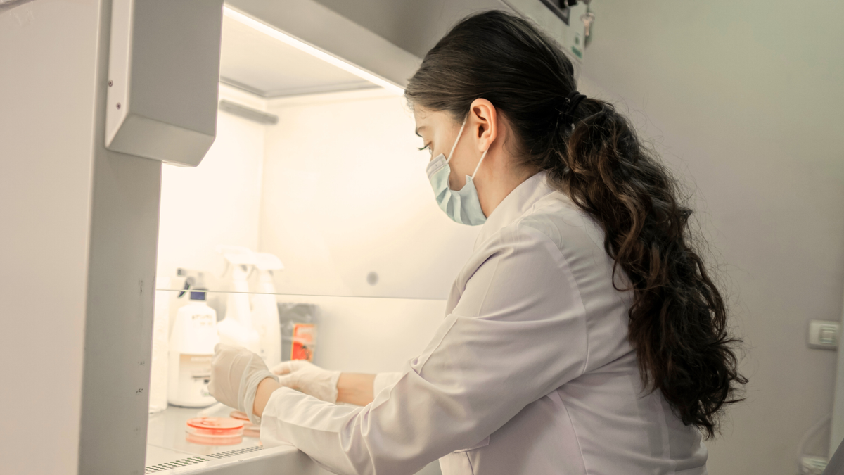 Person in a laboratory setting wearing a mask and gloves, working with equipment.